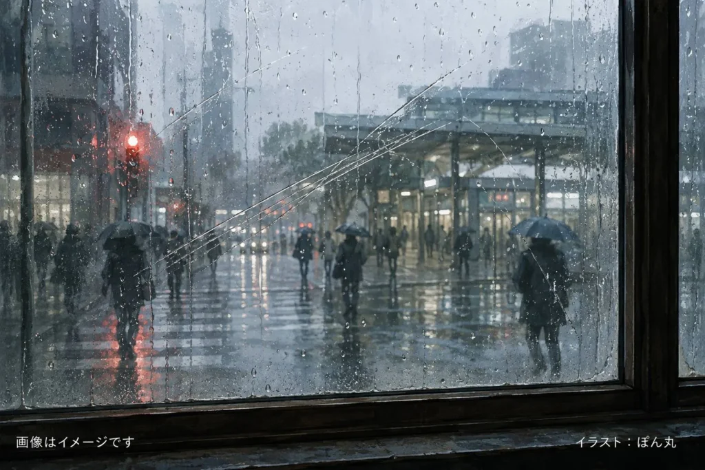 雨に濡れた都市の通りを、傷と雨跡の残る窓ガラス越しに眺めたイラスト。傘を差した人々がぽつぽつと駅へ向かい、濡れた路面には信号や街灯の光が長く反射している。全体に青灰色の静かな空気が漂い、朝の肌寒さと雨の日の孤独感が表現されている。画像左下に「画像はイメージです」、右下に「イラスト：ぽん丸」の文字入り。