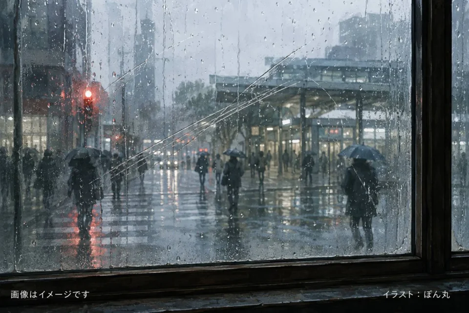 雨に濡れた都市の通りを、傷と雨跡の残る窓ガラス越しに眺めたイラスト。傘を差した人々がぽつぽつと駅へ向かい、濡れた路面には信号や街灯の光が長く反射している。全体に青灰色の静かな空気が漂い、朝の肌寒さと雨の日の孤独感が表現されている。画像左下に「画像はイメージです」、右下に「イラスト：ぽん丸」の文字入り。