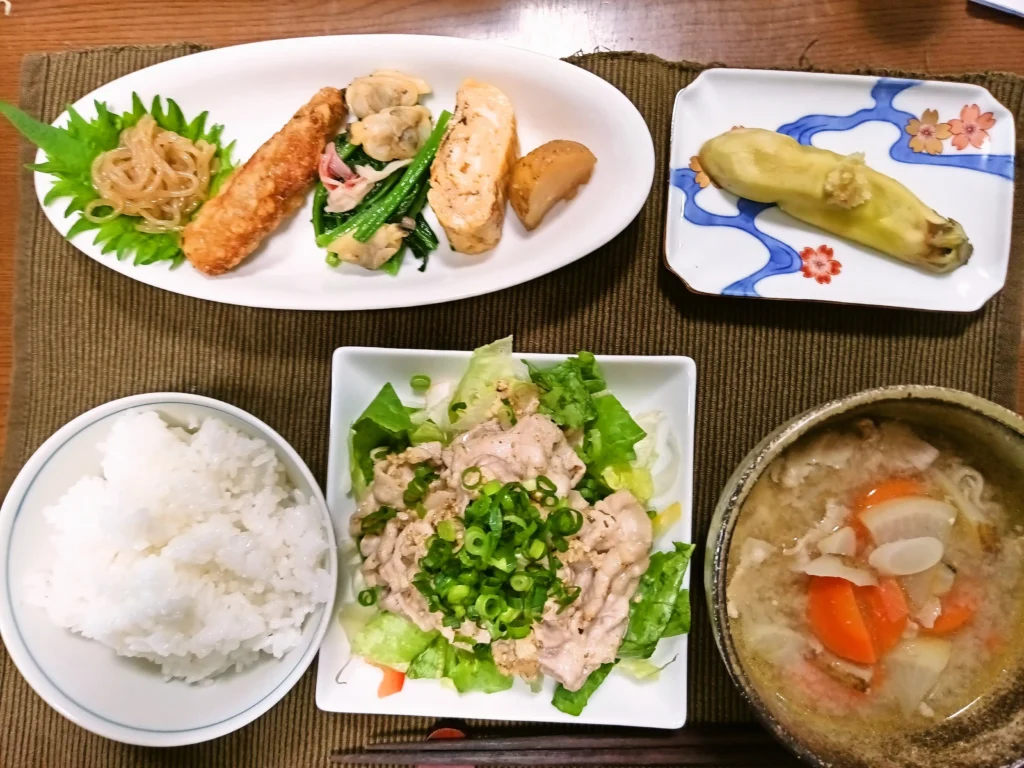 木のテーブルに並んだ和定食。中央に青ねぎをたっぷりのせた豚冷しゃぶサラダ。右下には具だくさんの豚汁、左下に白ご飯。上の白い皿には糸こんにゃく炒め、手羽中唐揚、あさりとほうれん草のバターソテー、玉子焼き、揚げじゃが煮が整然と並ぶ。右上の小皿には淡い黄色の冷やし茄子。全体に家庭料理らしい温かさがあり、茶色のランチョンマットと和柄の器が落ち着いた雰囲気を作っている。