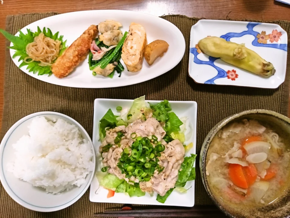 木のテーブルに並んだ和定食。中央に青ねぎをたっぷりのせた豚冷しゃぶサラダ。右下には具だくさんの豚汁、左下に白ご飯。上の白い皿には糸こんにゃく炒め、手羽中唐揚、あさりとほうれん草のバターソテー、玉子焼き、揚げじゃが煮が整然と並ぶ。右上の小皿には淡い黄色の冷やし茄子。全体に家庭料理らしい温かさがあり、茶色のランチョンマットと和柄の器が落ち着いた雰囲気を作っている。