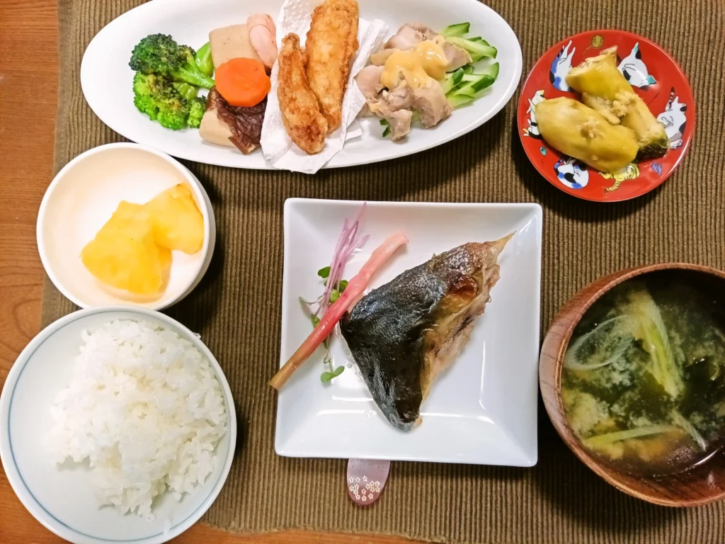 木目のテーブルに並んだ和定食。白い四角皿に焼いたぶりカマが一切れ、はじかみが添えられている。左下に白ごはん、右下にわかめとねぎのみそ汁。上の長皿には焼きブロッコリー、高野豆腐、にんじん、手羽中唐揚げ、蒸し鶏ときゅうりなどの副菜。小鉢にパイナップルが盛られている。全体に落ち着いた家庭料理の雰囲気。