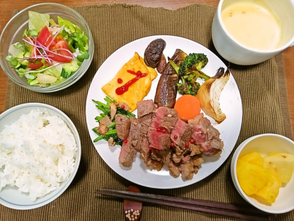 木のテーブルの上に並んだステーキ定食。白い皿にはミディアムレアの肩ロースステーキが盛られ、赤いピンクペッパーが散らされている。付け合わせに玉子焼き、焼き茄子、ブロッコリー、玉ねぎ、にんじん、しいたけ、青菜炒め。別皿で白ご飯、ガラス鉢のサラダ、コーンポタージュ、パイナップルも添えられている。全体に温かい家庭的な雰囲気。