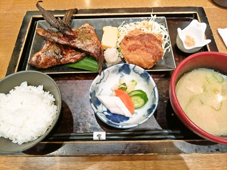 木製のお盆に載った和定食。左に白ご飯、右に豚汁。中央の長皿には、こんがり焼けた鯛カマが二切れと丸いエビカツが盛られ、千切りキャベツ、玉子焼き、大根おろしが添えられている。手前には胡瓜や大根、人参の漬物の小鉢。右上には小さな小鉢があり、全体に落ち着いた和食店の雰囲気。鯛カマの香ばしい焼き色が印象的な定食。