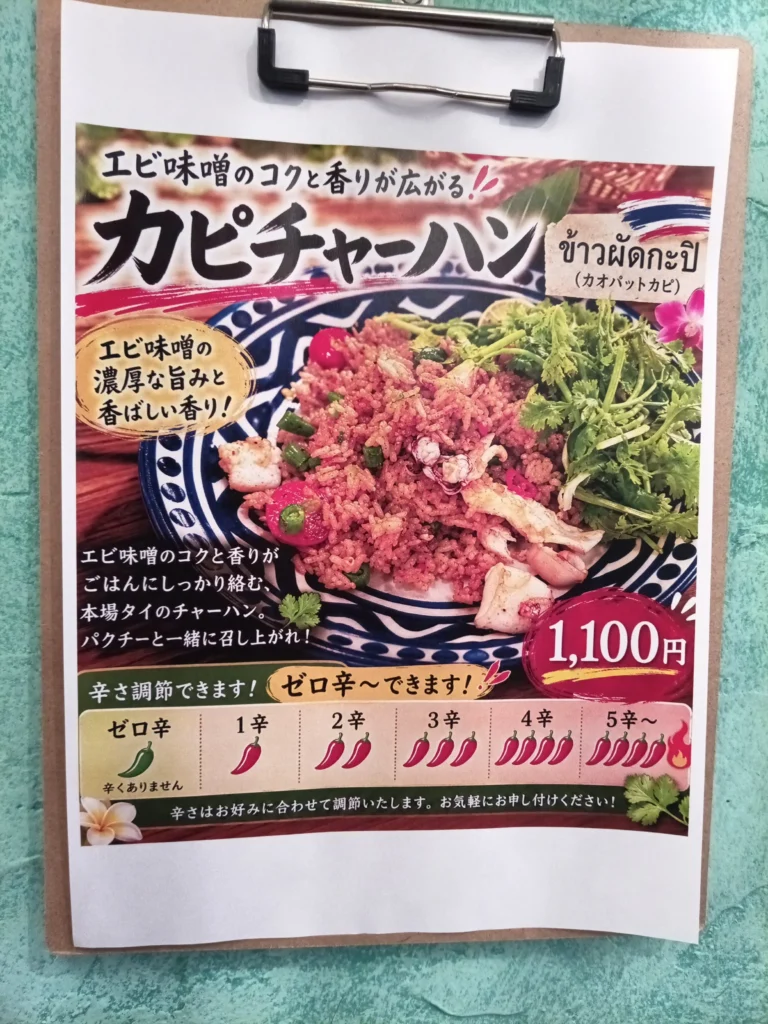 クリップボードに挟まれた週替わりメニューの案内。「カピチャーハン」と大きく書かれ、海老味噌のコクと香りを特徴とするタイ風チャーハンの写真が掲載されている。皿には赤みがかったチャーハン、イカや小海老、ミニトマト、青ねぎ、たっぷりのパクチーが盛られ、価格は1100円。下部には辛さをゼロ辛から五辛以上まで選べる表示があり、「ゼロ辛もできます」と案内されている。