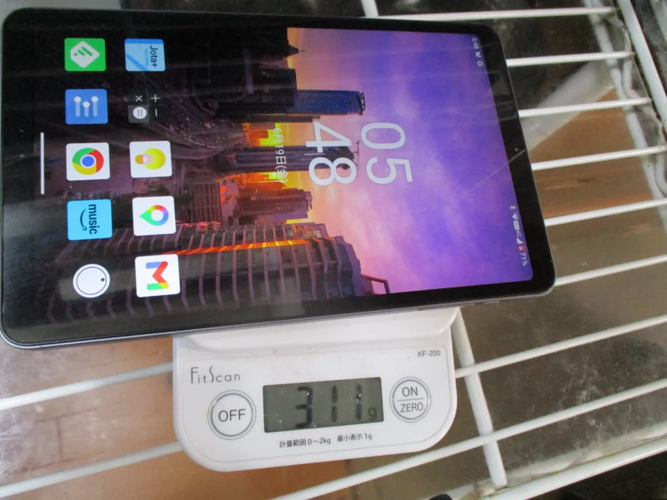 キッチンスケールの上に、8インチタブレットが斜めに載っている。画面には都市の壁紙とアプリのアイコンが表示され、スケールの液晶には「311g」と出ている。軽量を期待した新しいタブレットの重さを確かめている場面。