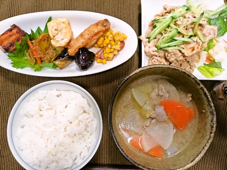 白いご飯と具だくさんの豚汁を中心にした家庭の昼食。別皿には手羽中の唐揚げ、玉子焼き、チャプチェ、揚げ茄子、コーンバター、焼き厚揚げ煮が彩りよく並び、もう一皿にはレタスときゅうりを添えた豚冷しゃぶサラダが盛られている。全体を上から撮影した、さっぱりしつつ品数豊富な定食風の一枚。