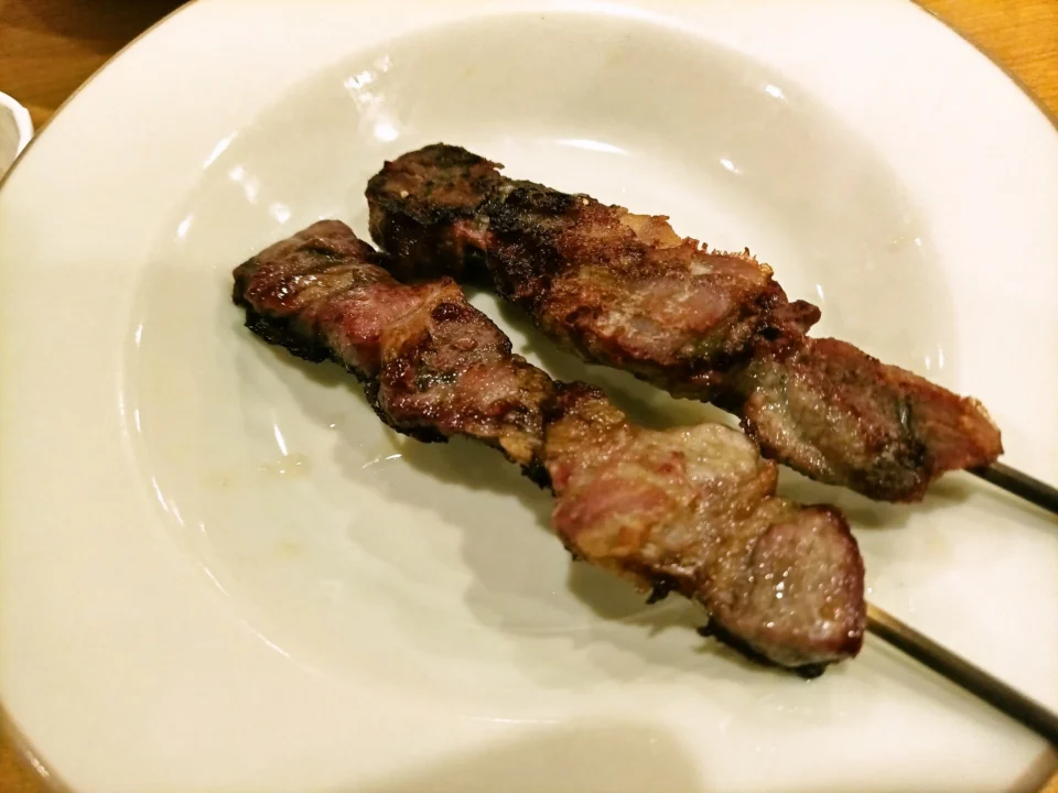 白い皿に、焼き色のついたアロスティチーニ(肉の串焼き)が二本のっている。表面は香ばしく焦げ目があり、脂がつやりと光っている。木目のテーブルの上で、温かい店内の光に照らされている。
