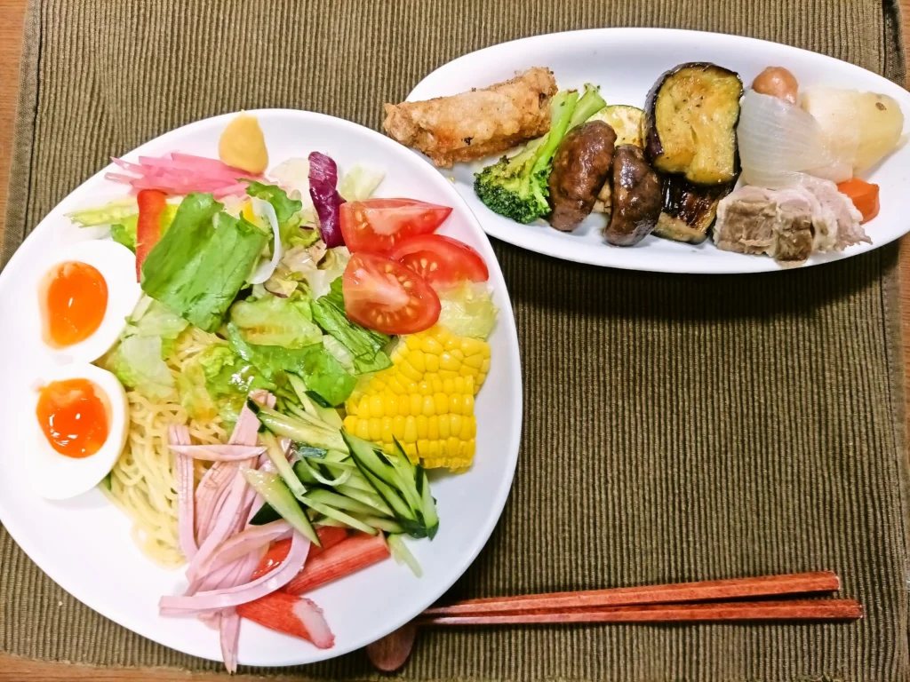 白い大皿に盛られた冷やし中華。麺の上には細切りハム、きゅうり、カニ風かまぼこ、レタス、トマト、赤パプリカ、コーン、半熟ゆで玉子が彩りよく並び、紅しょうがとからしが添えられている。隣の楕円皿には手羽中唐揚げ、ブロッコリー、椎茸、オリーブオイルで焼いた茄子とズッキーニ、ポトフの玉ねぎ、じゃがいも、人参、豚肉が盛られている。手前には木の箸が置かれ、家庭で丁寧に用意されたランチの雰囲気が伝わる。