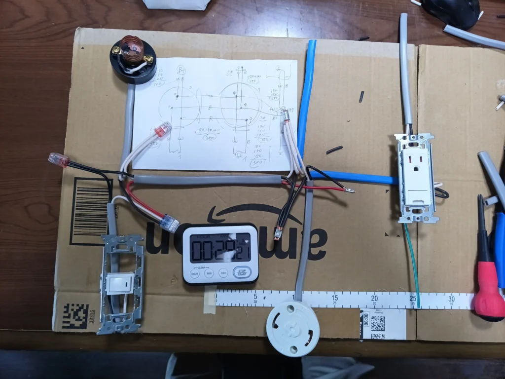 第二種電気工事士技能試験の練習中の作業台。段ボールの上に配線図のメモ、タイマー、ケーブル、スイッチ、コンセント、引掛シーリング、ランプレセプタクルなどが並んでいる。ケーブルの切れ端や工具も散らばり、銅線を少しも無駄にしたくない練習の真剣さが伝わる。