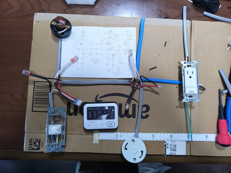 第二種電気工事士技能試験の練習中の作業台。段ボールの上に配線図のメモ、タイマー、ケーブル、スイッチ、コンセント、引掛シーリング、ランプレセプタクルなどが並んでいる。ケーブルの切れ端や工具も散らばり、銅線を少しも無駄にしたくない練習の真剣さが伝わる。