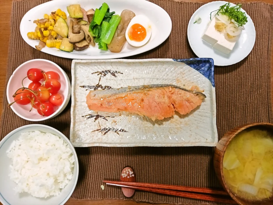 銀鮭西京焼きを中心にした和定食。長方形の皿に焼いた銀鮭が盛られ、左下に白ごはん、右下に玉ねぎ入りのみそ汁。左にはさくらんぼの小鉢。上の大皿には海老と椎茸とズッキーニのオリーブオイル炒め、小松菜のおひたし、こんにゃく、半熟の塩麹玉子、ベーコンとコーンのバター炒めが並ぶ。右上には冷や奴。全体に彩り豊かで、家庭料理らしい温かみのある食卓。