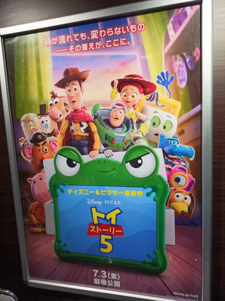 映画館の壁に掲示されたトイストーリー5のポスター。ウッディ、ジェシー、バズたち玩具が、緑色のカエル型タブレットの後ろから不安そうに画面を見つめている。上部には「時が流れても、変わらないもの。その答えが、ここに。」、下部には「7.3劇場公開」と記されている。